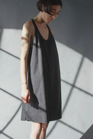 Scoop neck cocoon shaped effortless dress, knee length made of crisp black cotton poplin. Model has short straight black hair, and tatoos on her arms. The background is grey with angular box shadows, high contrast between the shadows and grey. Her arms are resting at her side and she is looking down, hair kinda falling in her face. 